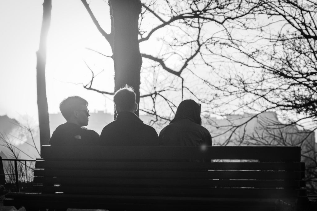 Three people sitting on a park bench, silhouetted by the sunset. A contemplative moment captured in black and white.